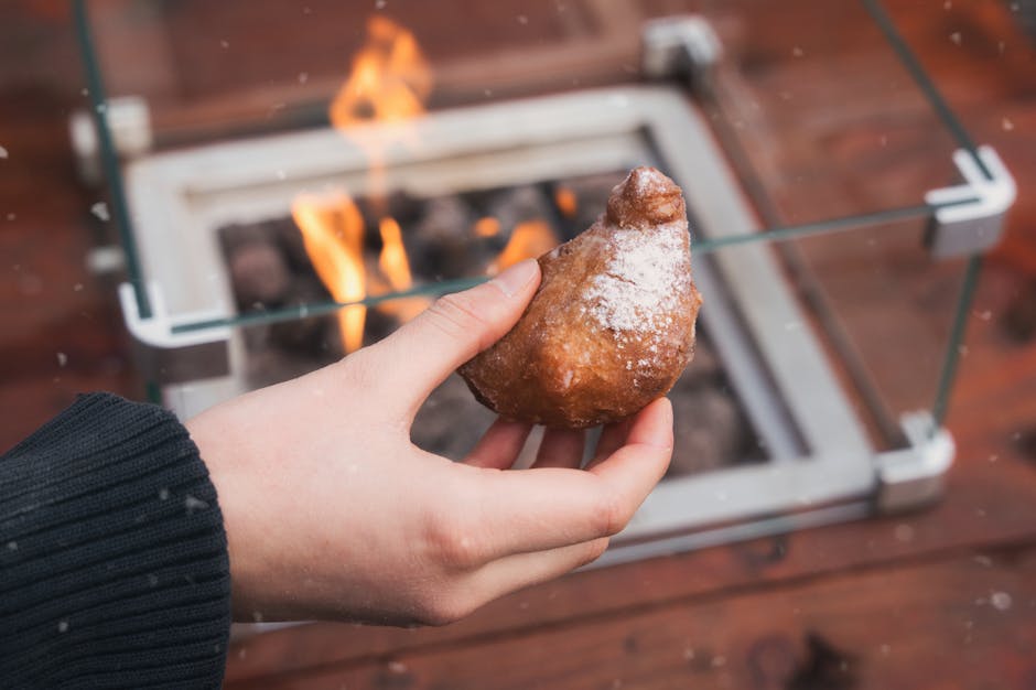 A hand holding a traditional Dutch oliebol dusted with powdered sugar, warm by an outdoor fireplace.