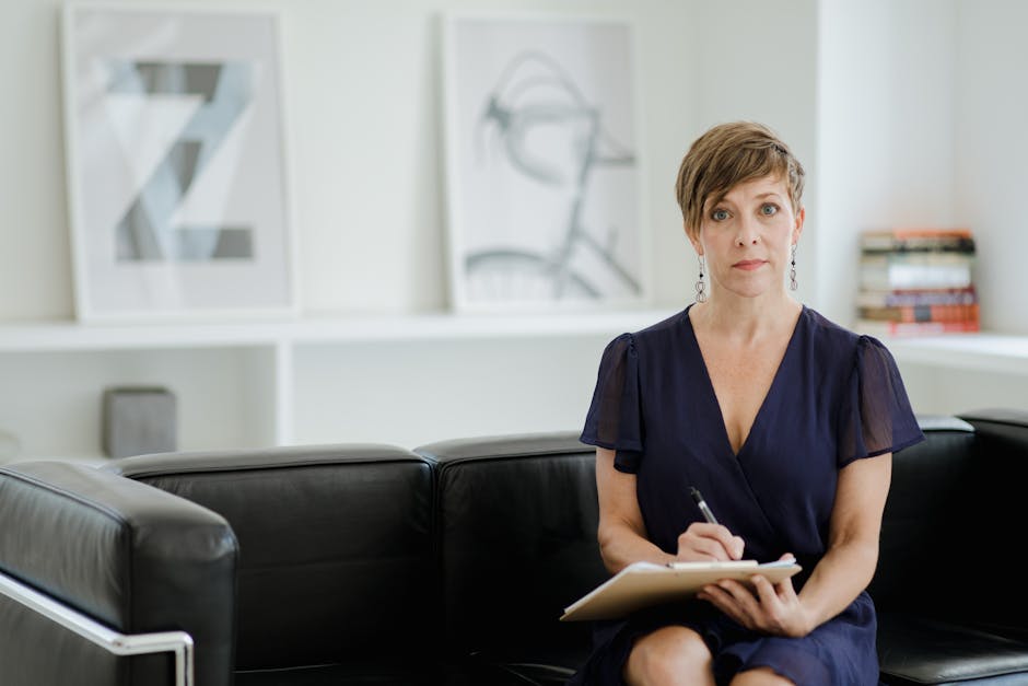 Professional woman sitting on a black couch, writing on a clipboard in a modern, bright office setting.