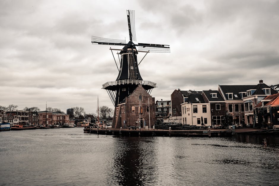 Scenic view of a traditional Dutch windmill by a river with urban buildings under cloudy skies.