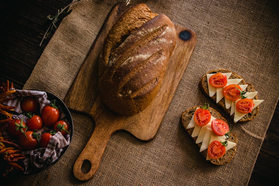 Artisan bread on a cutting board with cheese and tomato slices on burlap cloth.